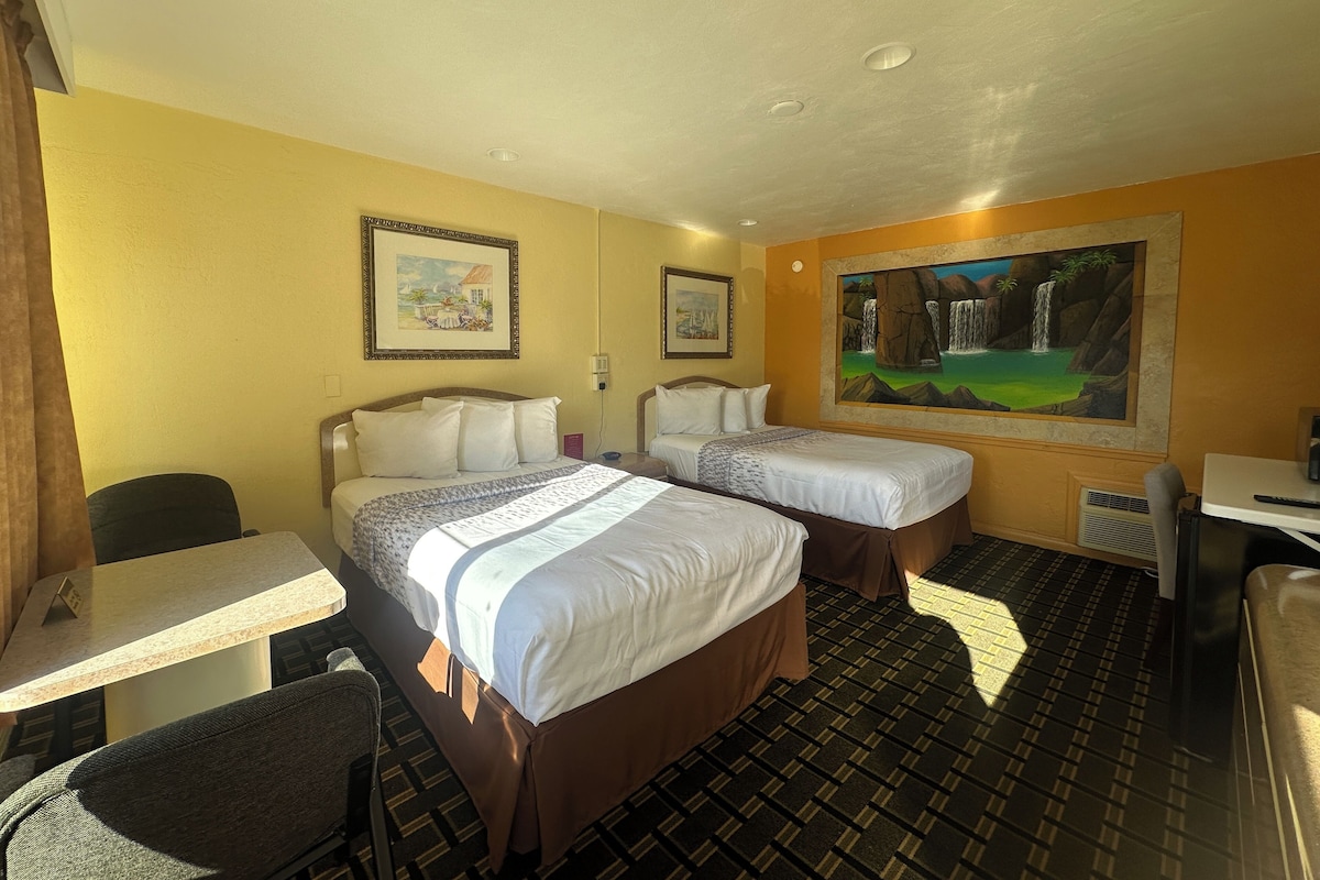 A hotel room with two beds, a desk, a chair, and a large painting on the wall.