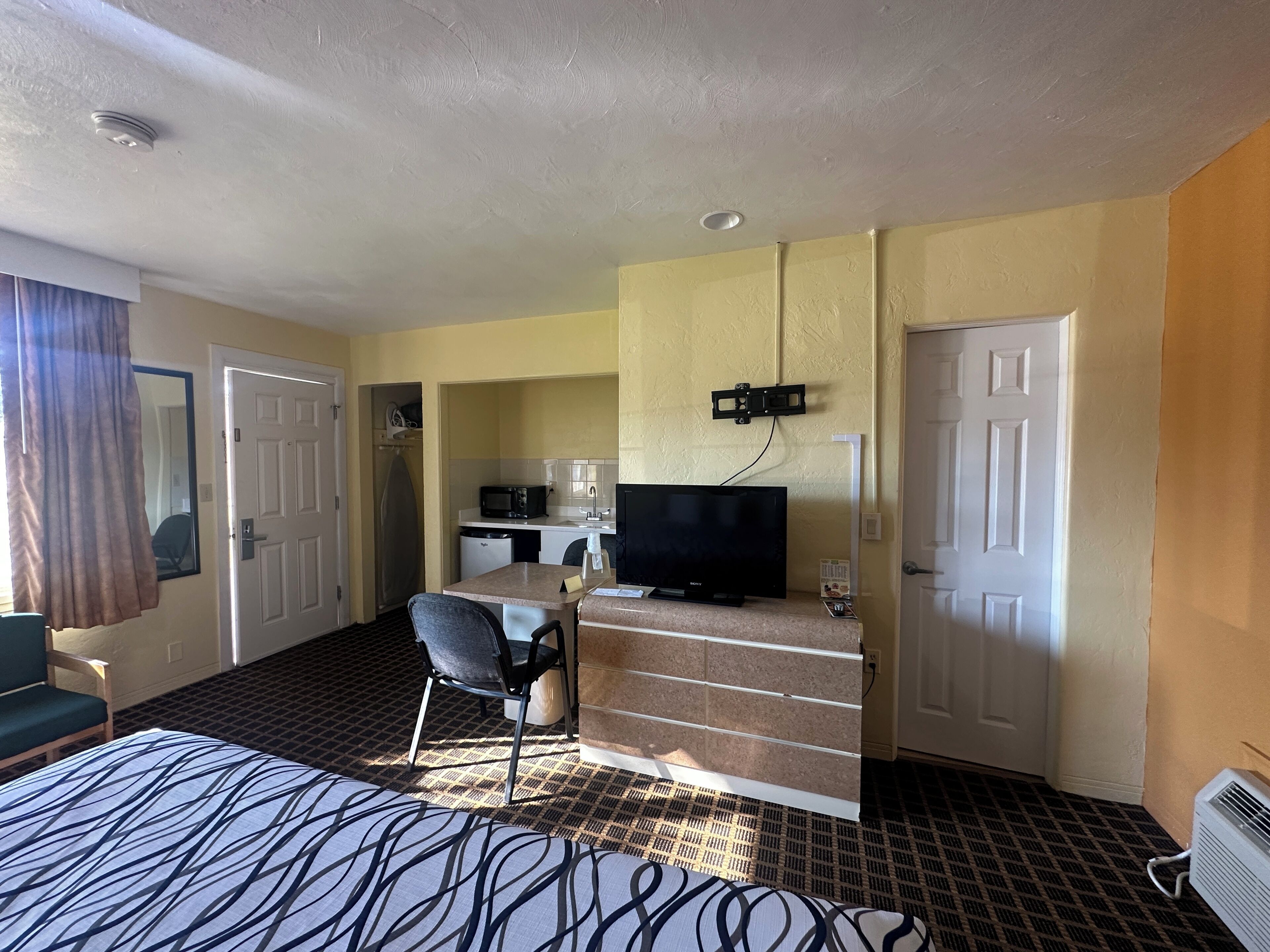 A hotel room with a bed, a desk with a television, a chair, and a kitchenette.