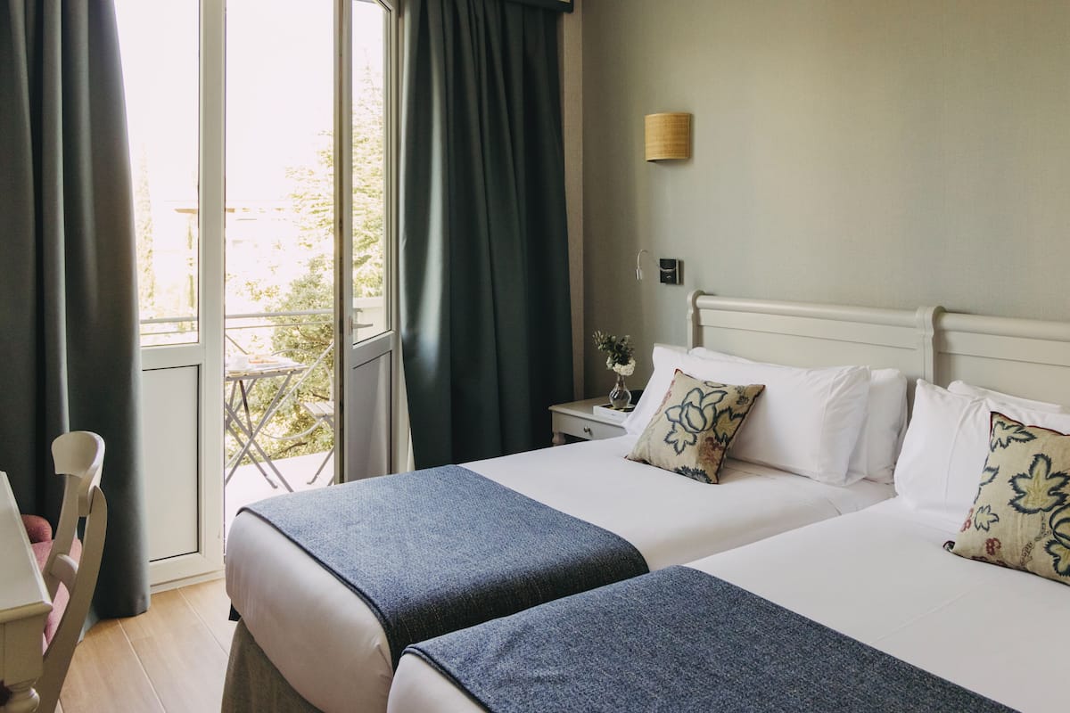 Een hotelkamer met twee bedden, een raam met gordijnen, een kleine tafel met een vaas en een wandlamp.