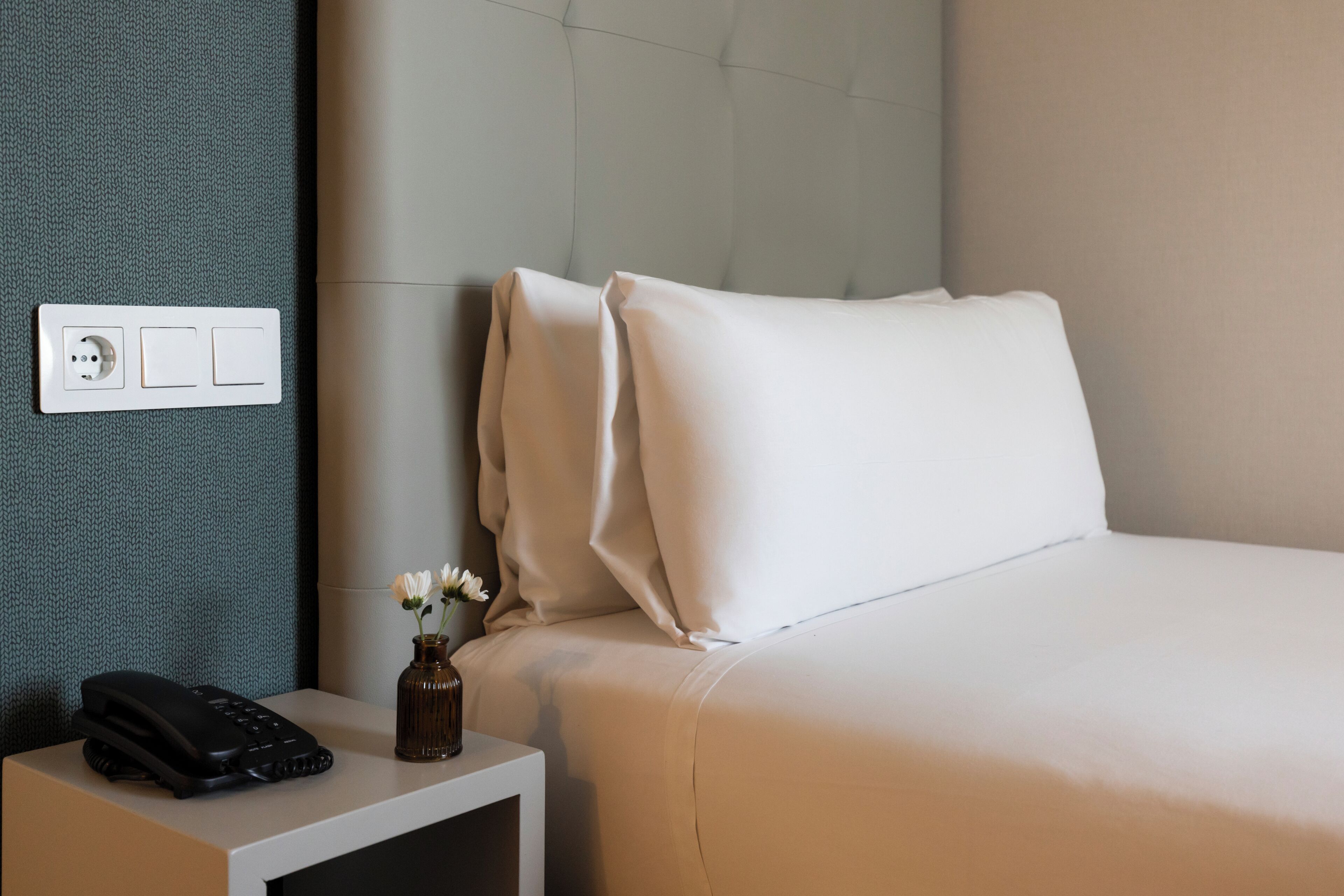 Et hotelværelse med en seng, et natbord med telefon og vase, samt en stikkontakt.