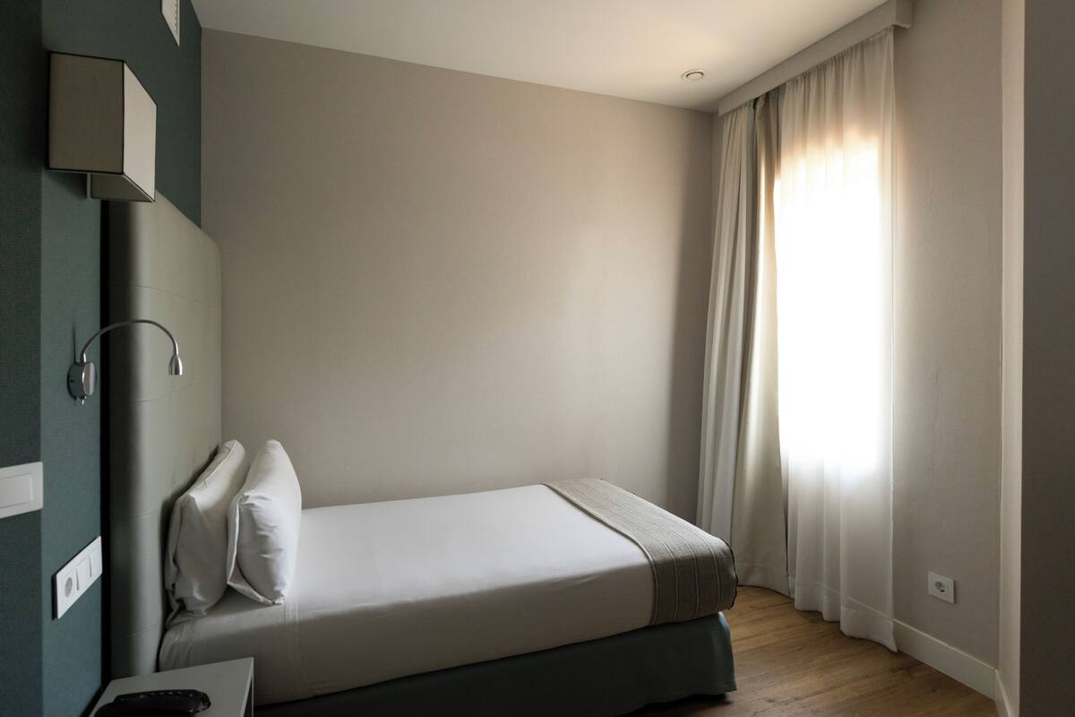 Een eenpersoonsbed in een kamer met een houten vloer, een nachtkastje met een lamp en een raam met gordijnen.