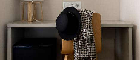 Una habitación con una televisión de pantalla plana montada en la pared, un escritorio con una lámpara, un mueble negro pequeño y una silla con un sombrero negro encima.