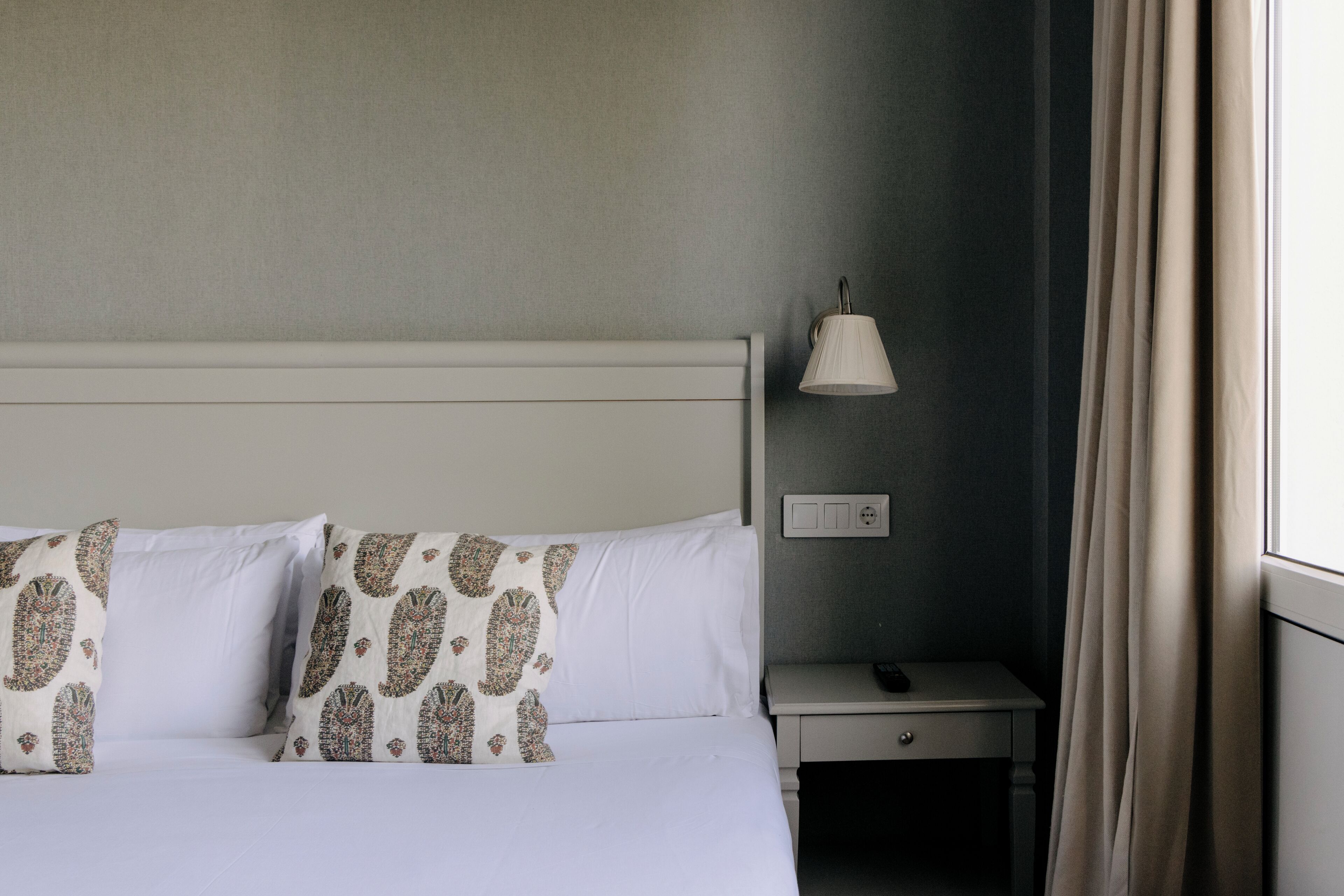A hotel room with a bed, two patterned pillows, a nightstand with a lamp, and a window with curtains.