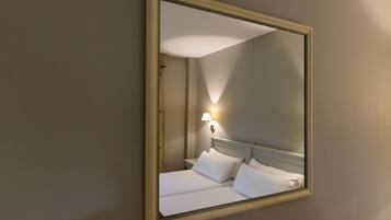 Habitación de hotel con un espejo grande, una cama con ropa blanca y una mesita de noche montada en la pared con una lámpara.