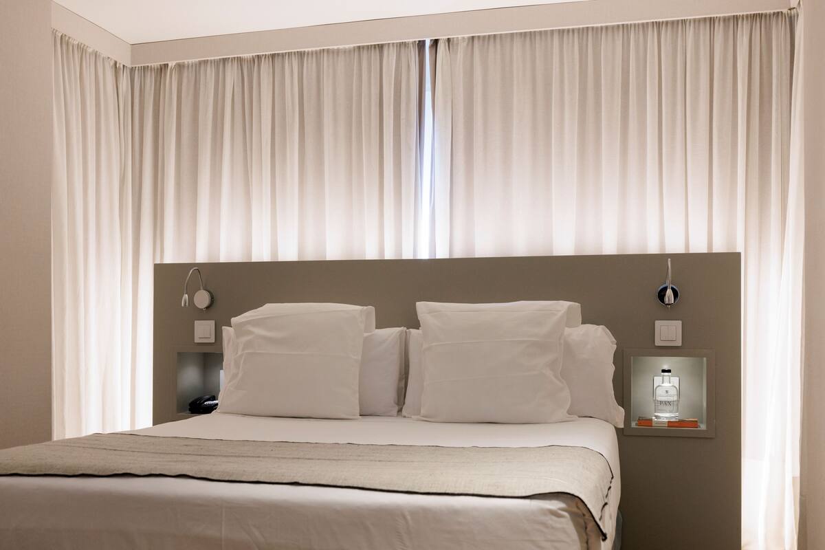 Een net opgemaakt bed met witte beddengoed en kussens, een nachtkastje met een lamp, en een op de muur bevestigde thermostaat.