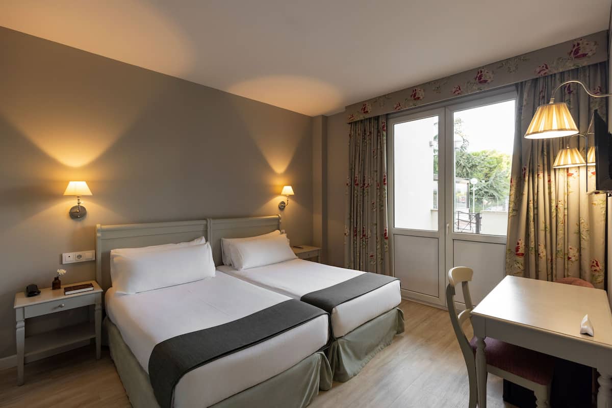 Een hotelkamer met twee bedden, een bureau en een stoel. Er staan lampen op de nachtkastjes en een raam met gordijnen.