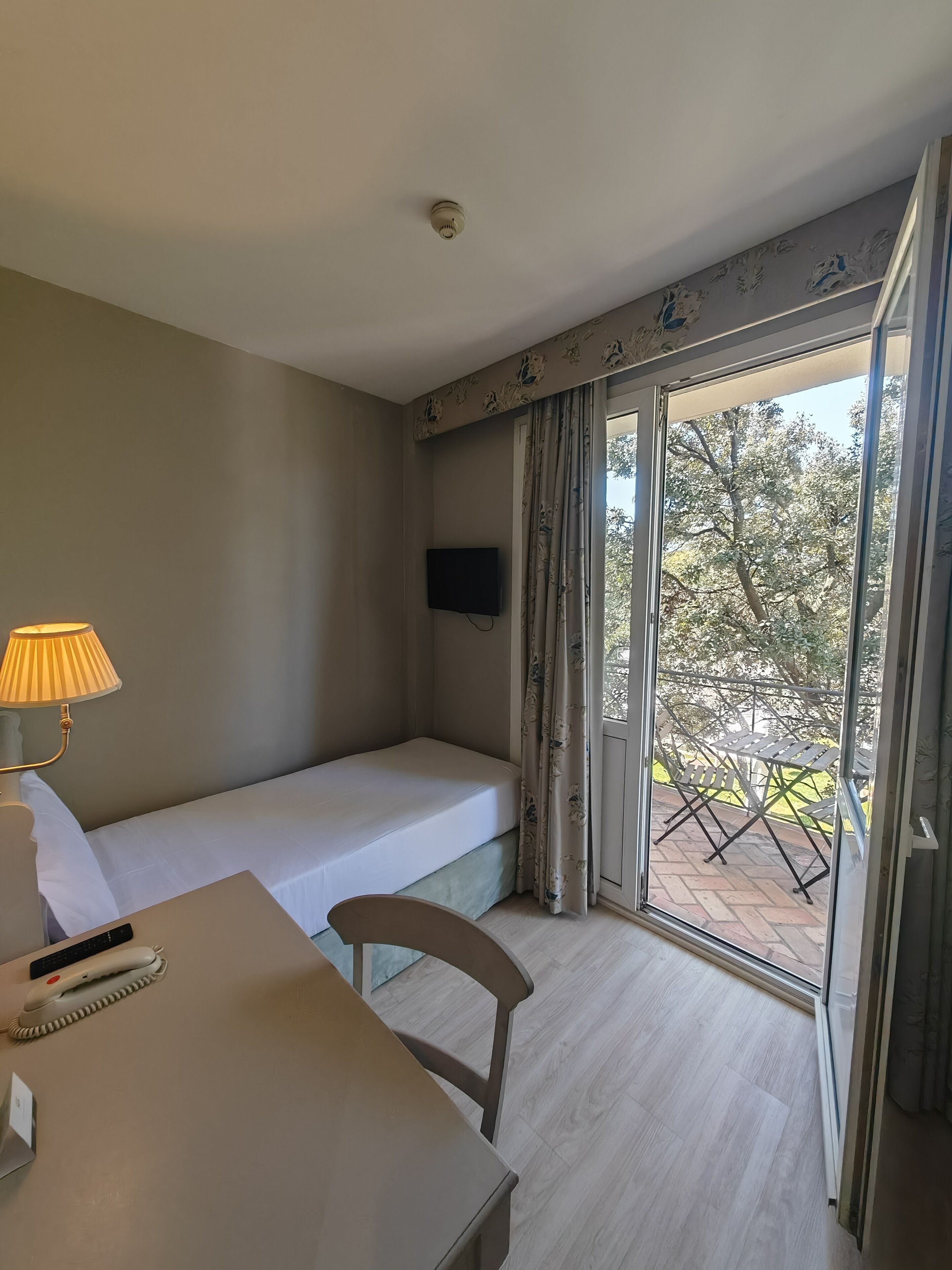 A hotel room with a single bed, a desk with a lamp, a chair, a TV mounted on the wall, and a sliding glass door leading to an outdoor area with tables and chairs.