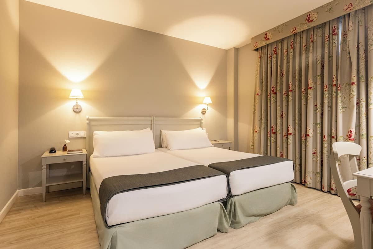Een tweepersoonsbed met twee nachtkastjes, een bureau en een stoel in een kamer met bloemen gordijnen en wandlampen.