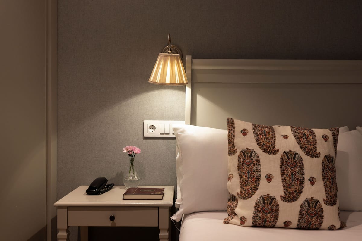 Een netjes opgemaakt hotelkamer met een bed, een bedlampje, een nachtkastje met een telefoon en een boek, en een vaas met bloemen.