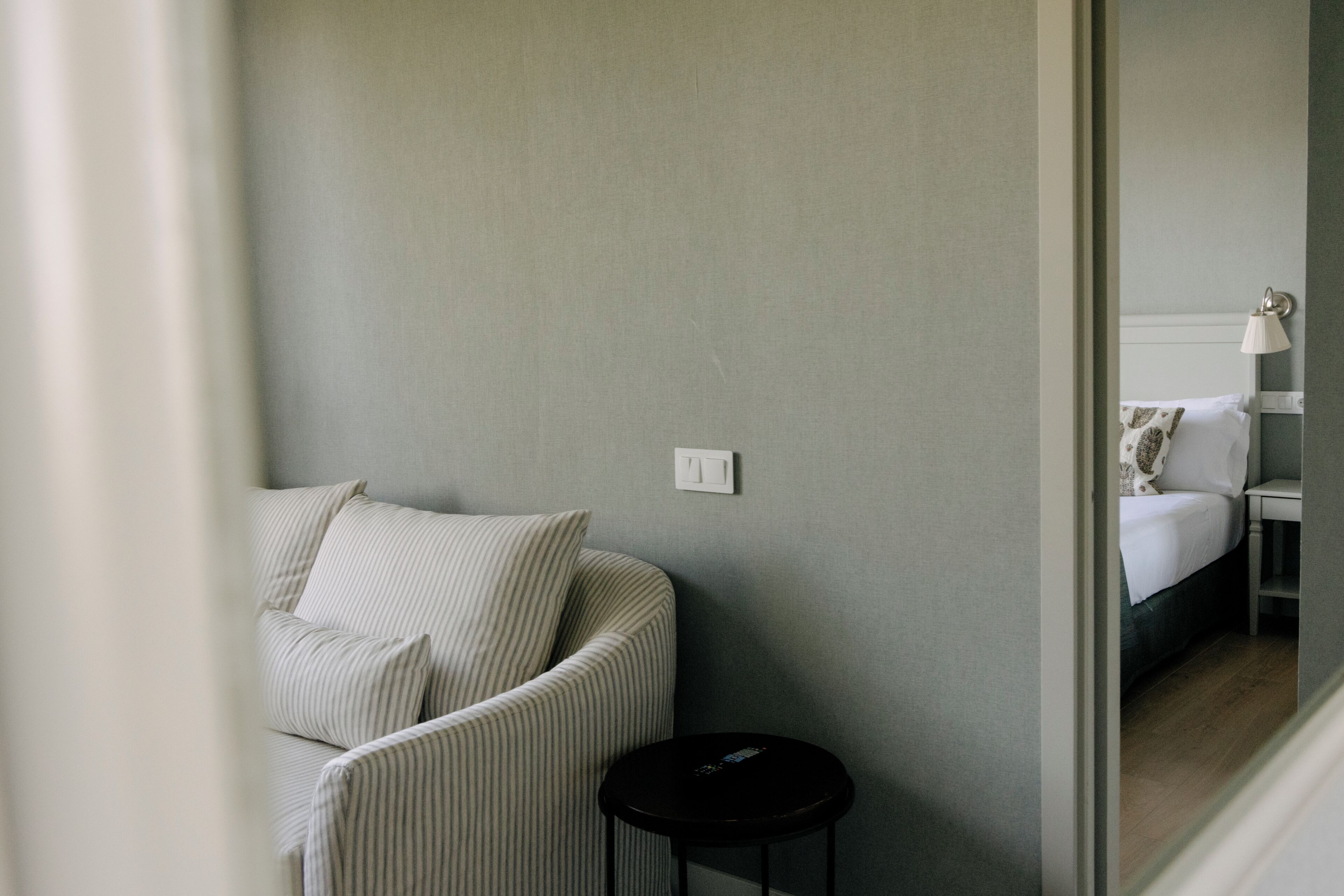 A room with a striped sofa, a small black stool, and a bed with a white headboard and patterned pillows.