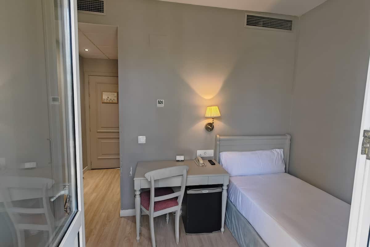 Een kleine hotelkamer met een enkel bed, een bureau met stoel, een nachtkastje en een lamp.