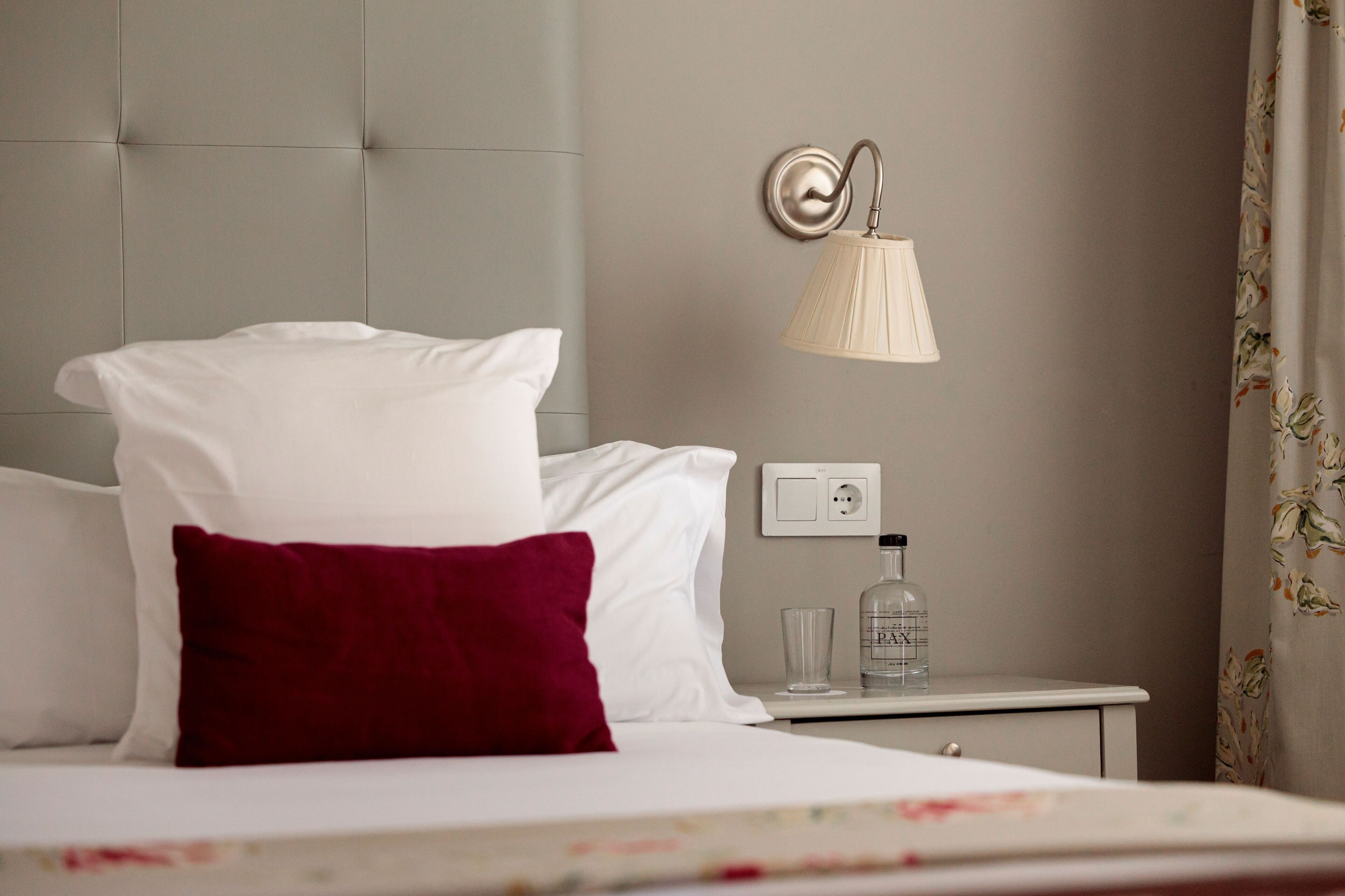 A hotel room with a bed, white and red pillows, a nightstand with a lamp, a bottle, and a glass.