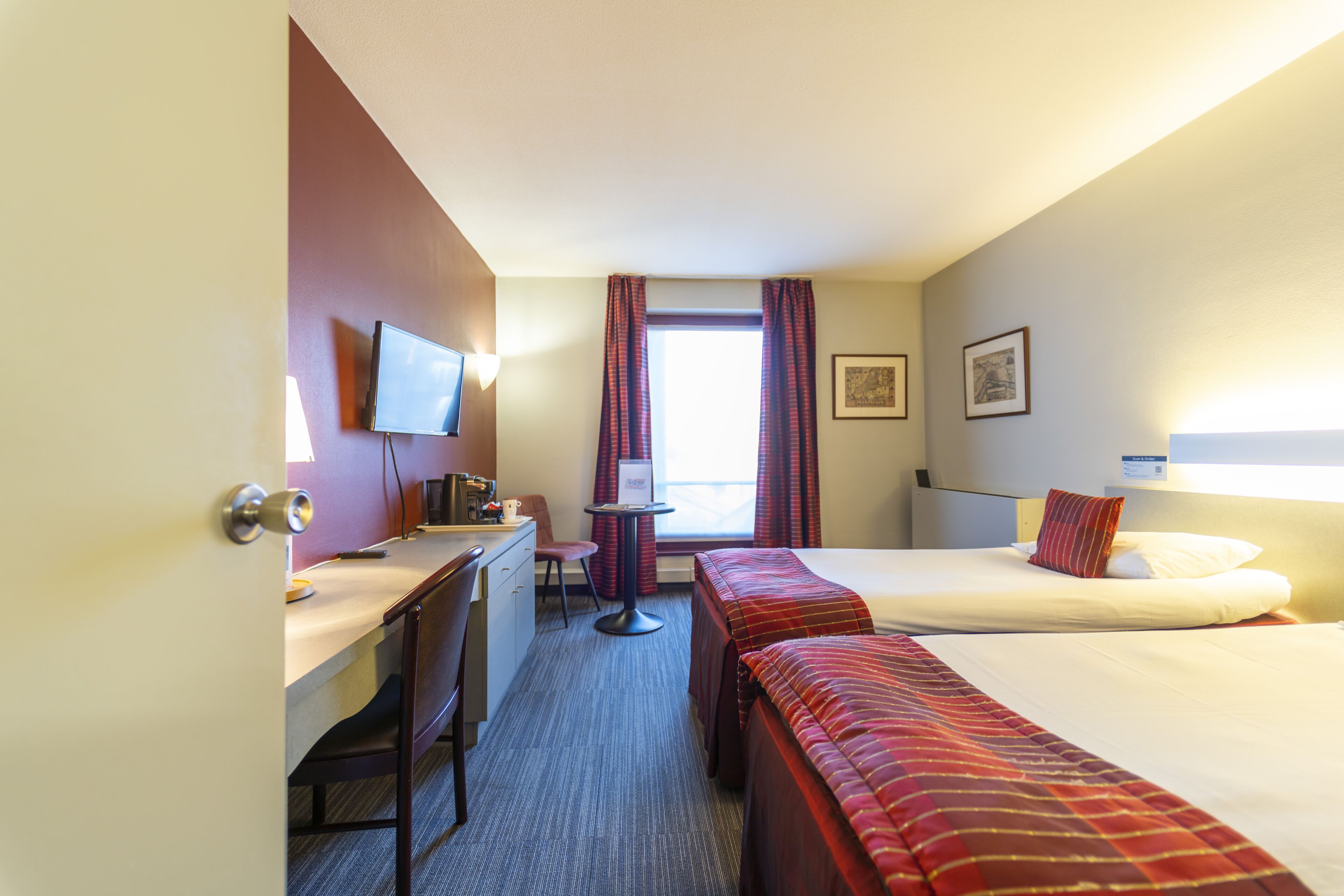 A hotel room with a desk, two beds, a TV, a window with curtains, and a framed picture on the wall.