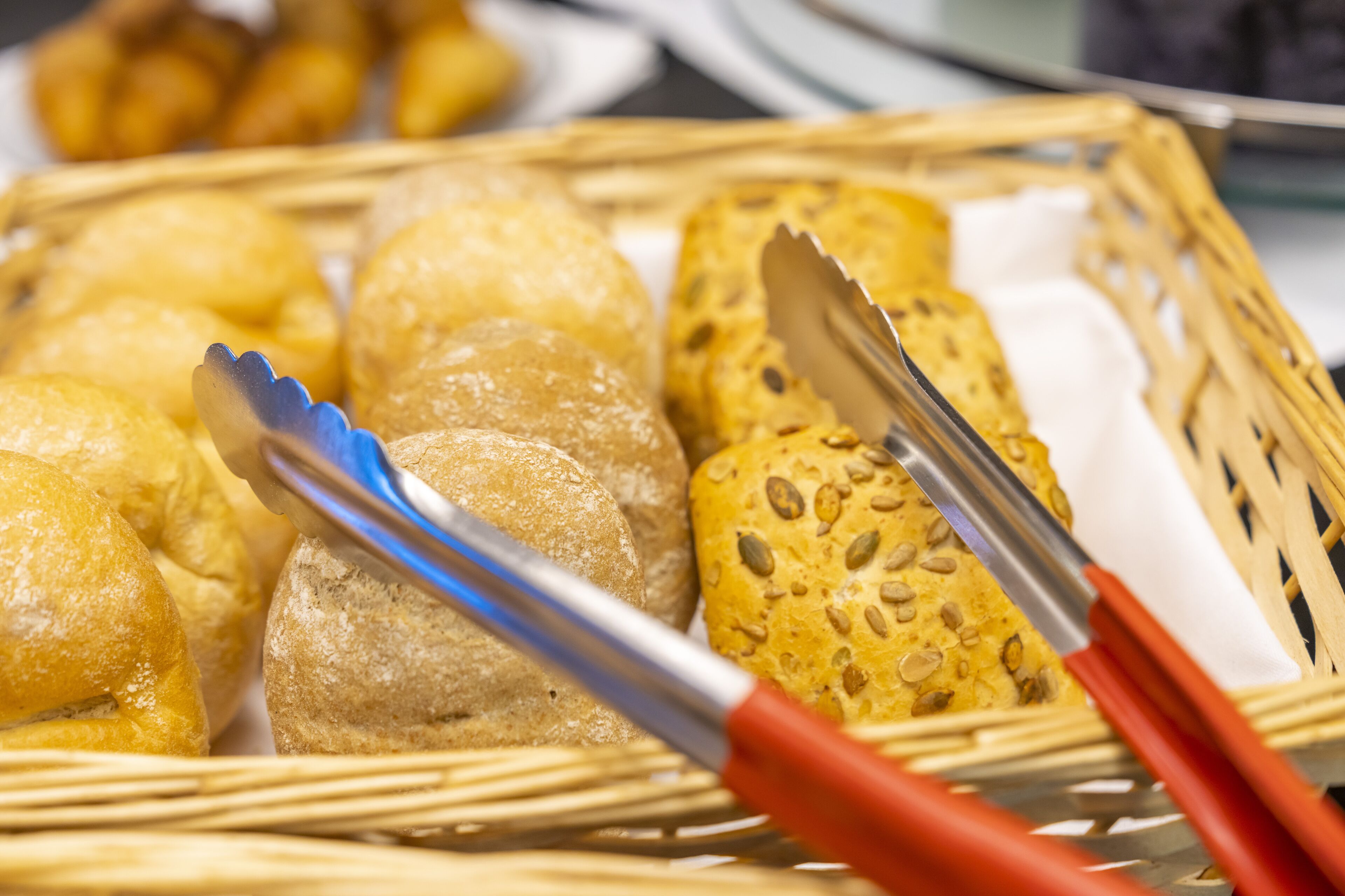 A basket of various breads and rolls, with tongs resting on top.