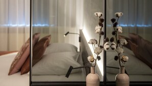 Ett modernt hotellrum med en stor spegel som reflekterar en säng och ett fönster med tunna, genomskinliga gardiner. På ett träbord står två vaser med torkade blommor.
