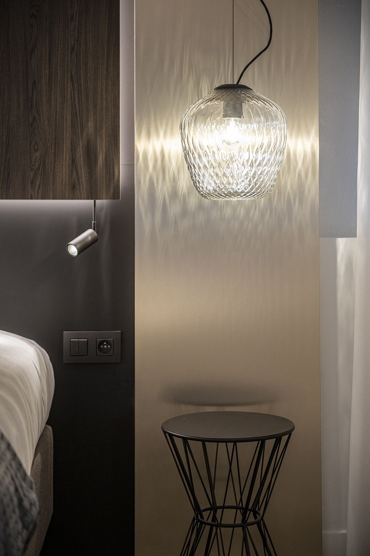 A modern bedroom with a wooden headboard, a bedside lamp, a wall-mounted light fixture, and a small round table.