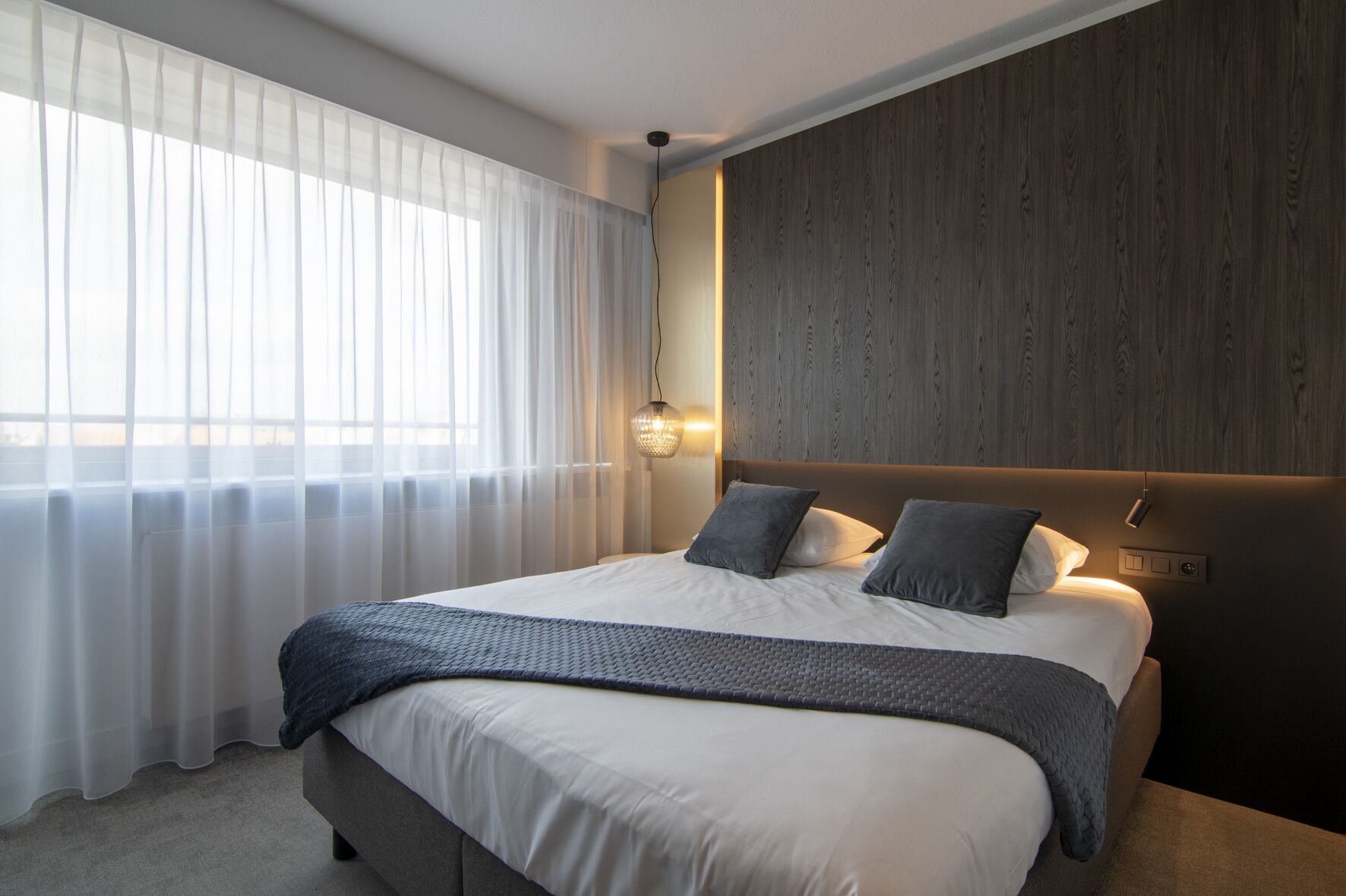 Een moderne slaapkamer met een groot bed, lichte gordijnen, een houten hoofdeinde en een wandlamp.