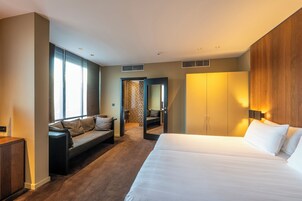 Una habitación de hotel moderna con una cama grande, un sofá, un armario y un baño visible a través de una puerta abierta.