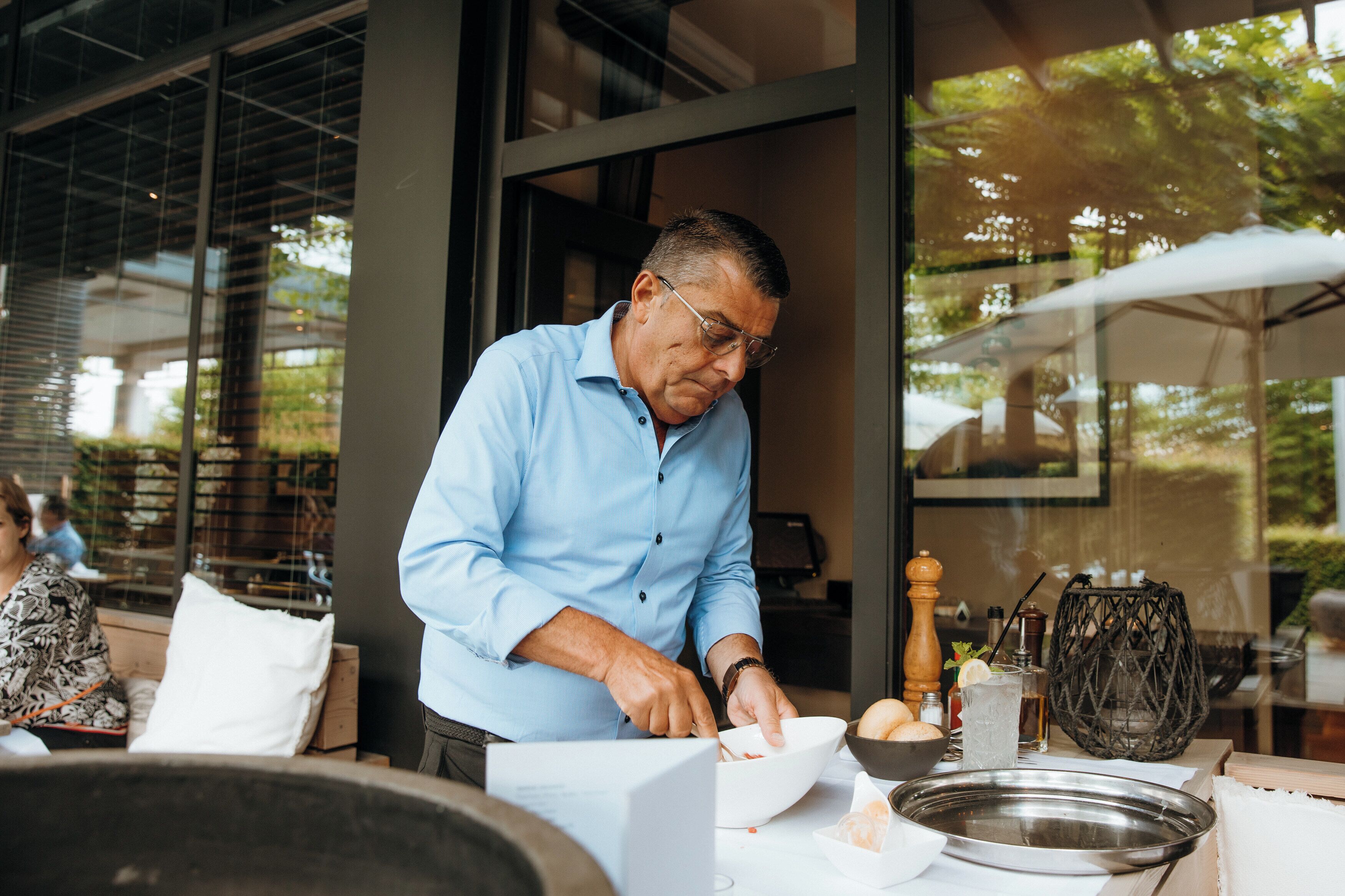 A person preparing food at an outdoor dining area with a modern architectural setting, including large windows and a view of greenery.