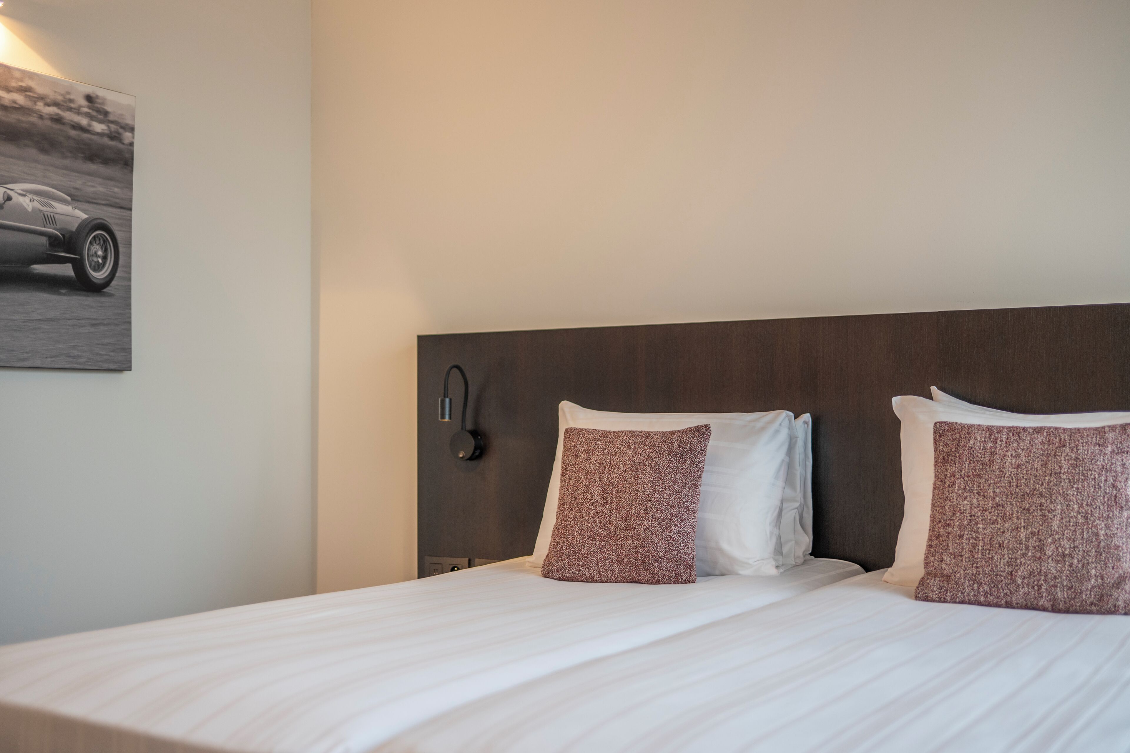 A neatly made bed with white linens and two patterned pillows, a dark headboard, and a wall-mounted lamp.