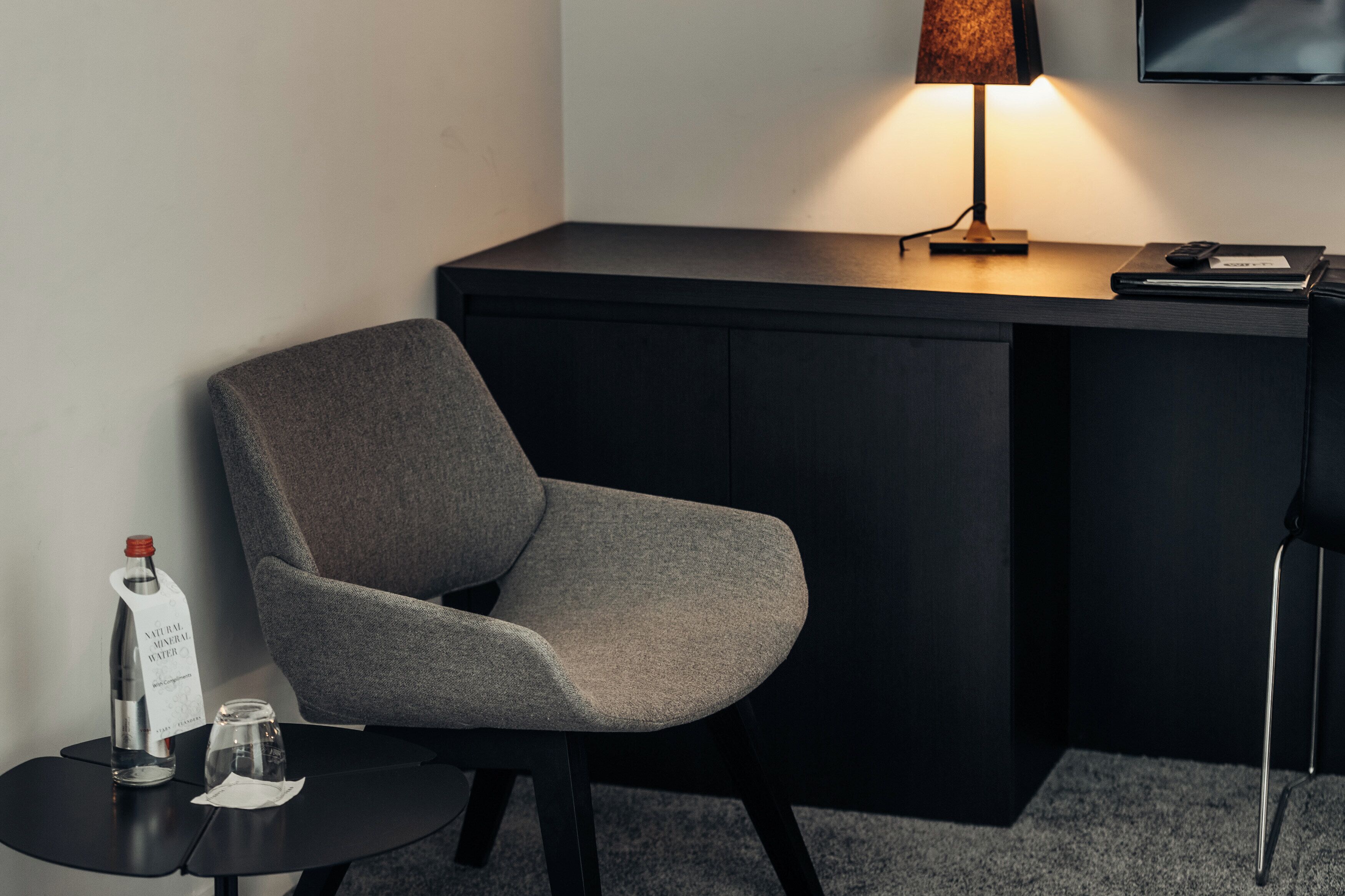 A modern room with a grey upholstered chair, a small round table with a bottle and glass, a desk with a lamp, and a flat-screen TV mounted on the wall.