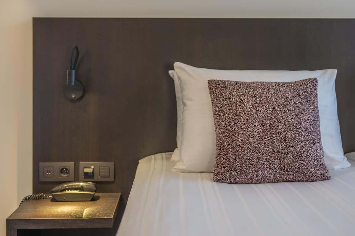 A neatly made bed with a white pillow and a patterned pillow, a bedside telephone, and a wall-mounted light fixture.