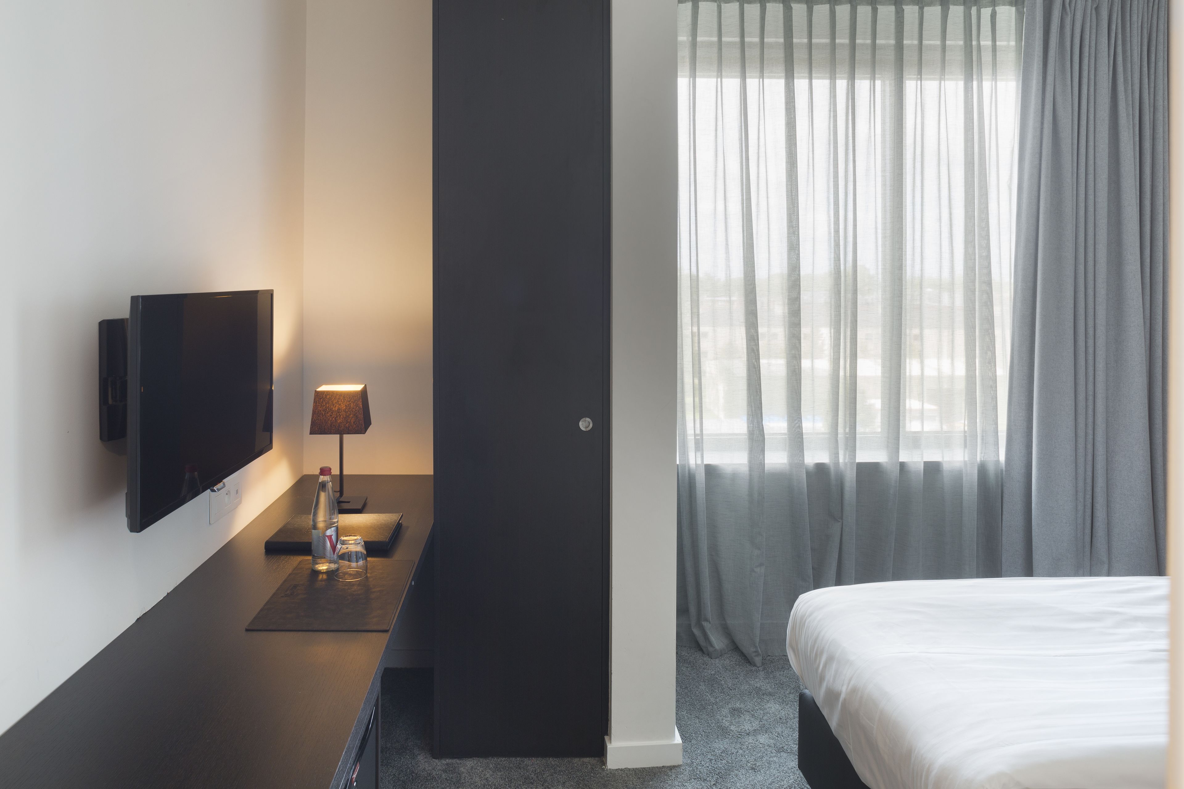 A hotel room with a bed, a flat-screen TV mounted on the wall, a bedside table with a lamp, and a window with sheer curtains.