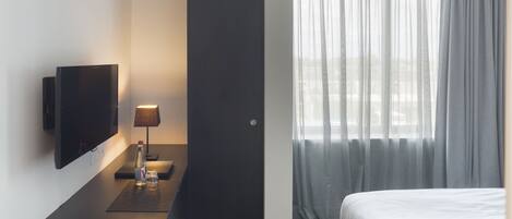 A hotel room with a bed, a flat-screen TV mounted on the wall, a bedside table with a lamp, and a window with sheer curtains.