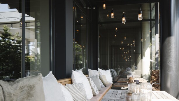 Un long espace étroit aménagé en coin salon, avec un plancher en bois, des bancs rembourrés et une rangée de lampes suspendues.