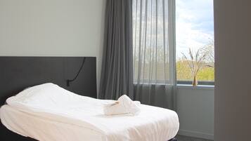 A neatly made bed with white linens and a folded towel, a dark headboard, and a window with sheer curtains and a view of trees.