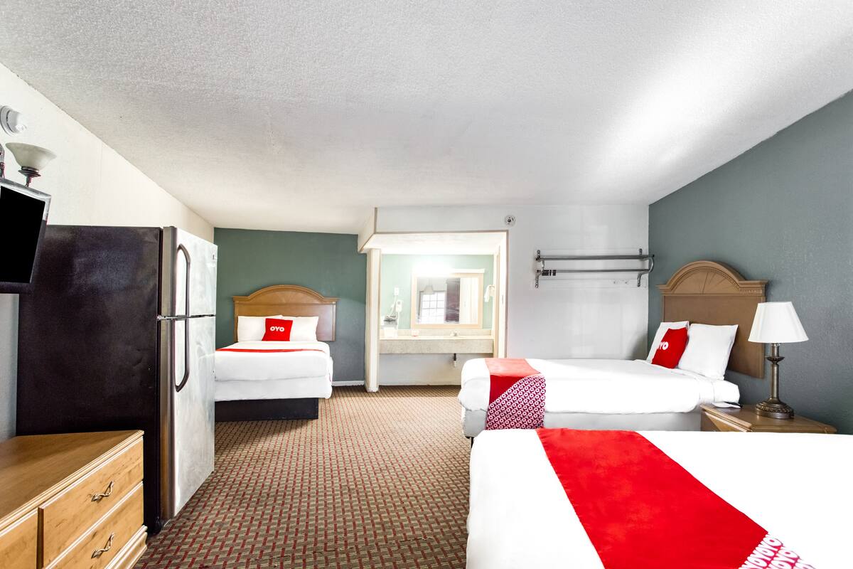 A hotel room with two beds, a refrigerator, a wooden dresser, a bathroom with a mirror, and a nightstand with a lamp.