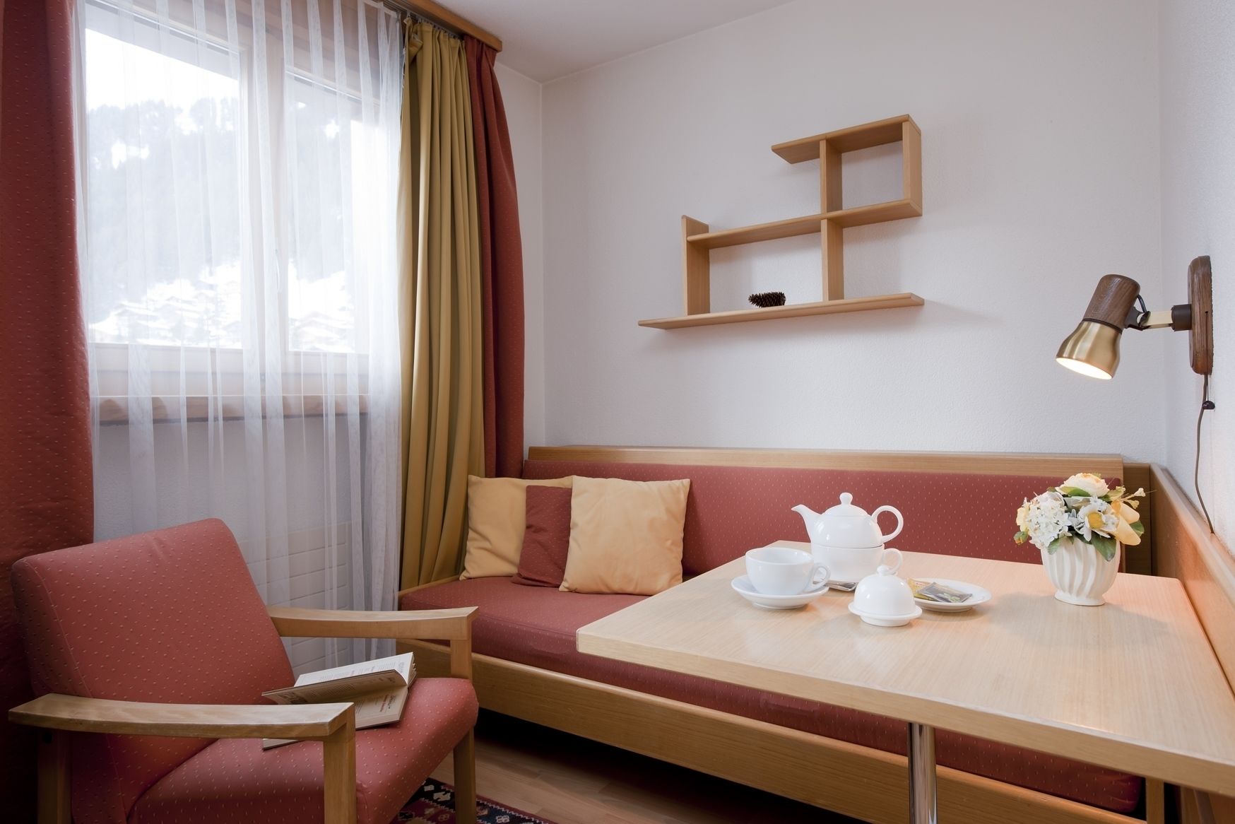 A cozy hotel room with a wooden bench, a small table with a teapot and cups, a vase with flowers, and a wall-mounted shelf.