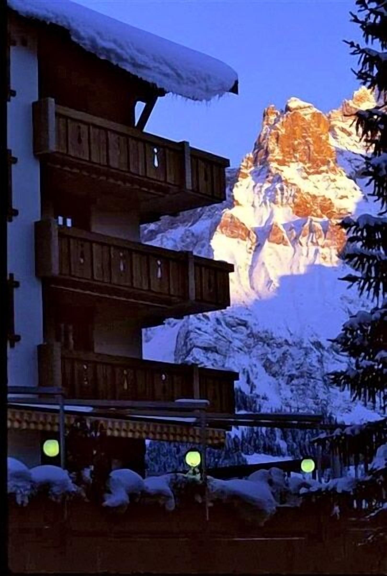 Ein mehrstöckiges Gebäude mit hölzernen Balkonen, umgeben von schneebedeckten Bergen und Bäumen.