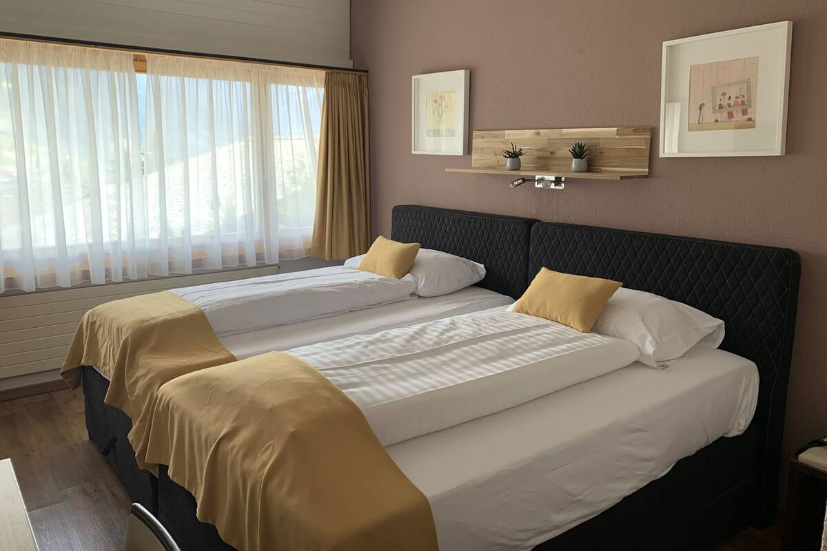 A double bed with two pillows, a bedside table, a window with sheer curtains, and two framed pictures on the wall.