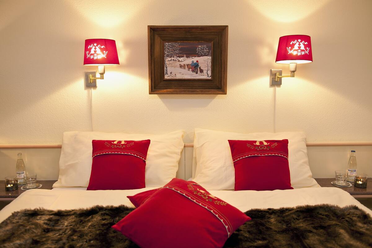 A hotel room with a bed, two red pillows, a fur throw, two nightstands with glasses and bottles, and two wall-mounted lamps with a painting above the bed.