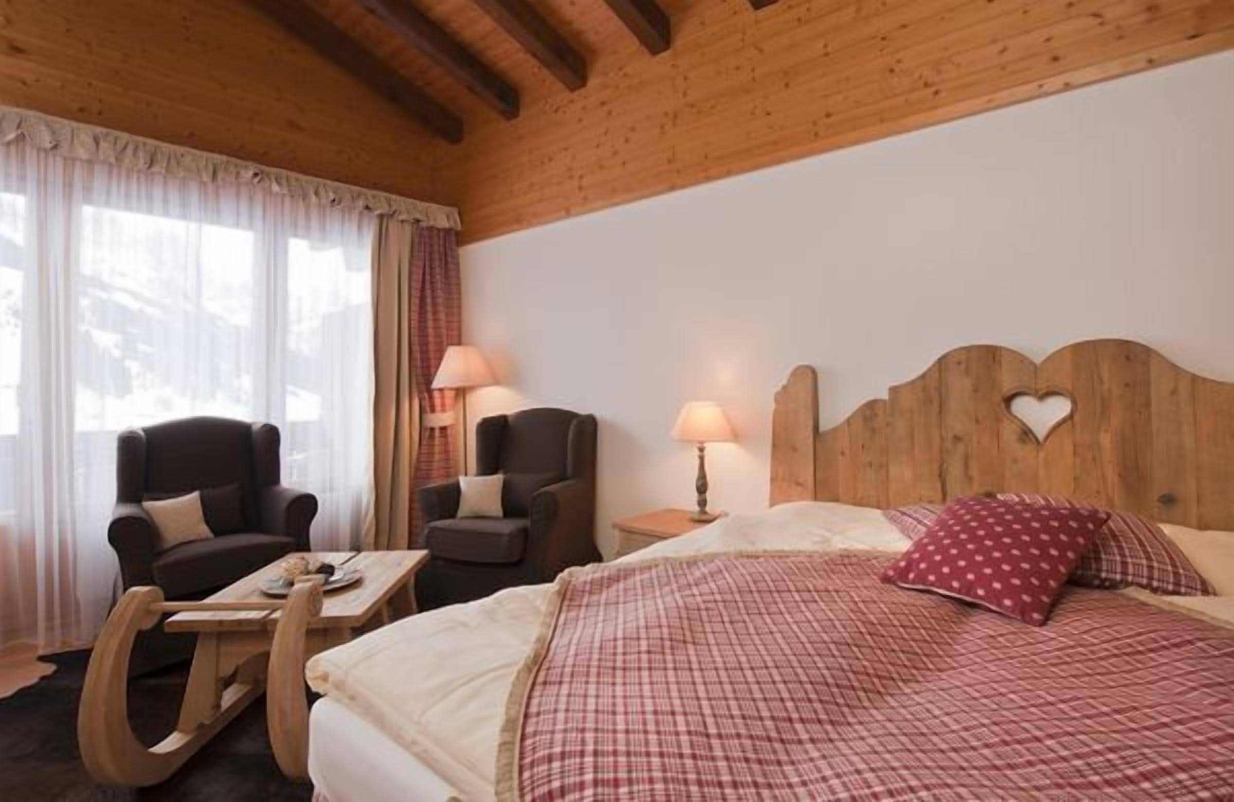 A cozy room with a wooden ceiling, a bed with a red checkered bedspread, two armchairs, a wooden coffee table, and a nightstand with lamps.
