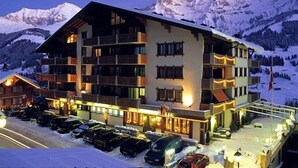 Un hotel de varios pisos con balcones, una pista de esquí al fondo y un estacionamiento con coches.