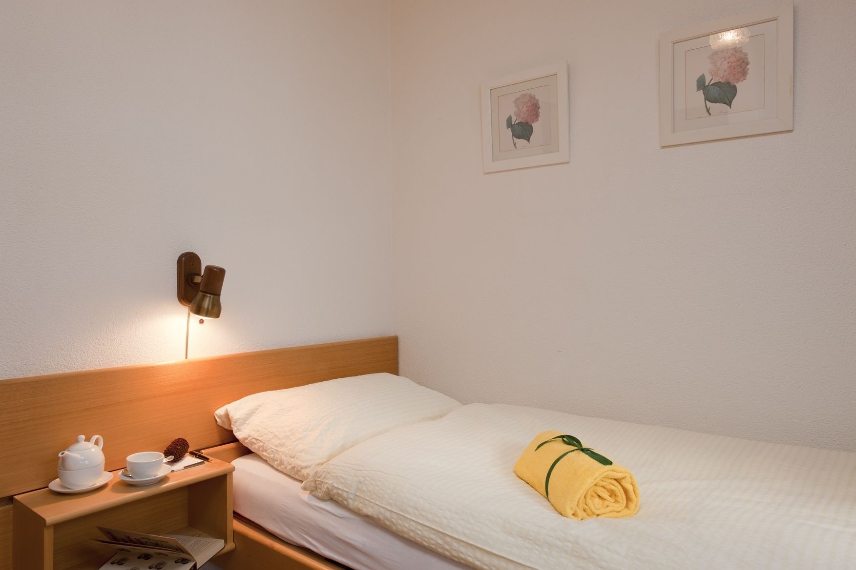 Habitación de hotel con cabecera de madera, mesita de noche con juego de té, toalla enrollada y dos cuadros de flores en la pared.