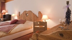 Un dormitorio acogedor con cabecera de madera, dos lámparas de noche, una cama con manta escocesa, una silla y una mesa con copas de vino y un ramo de flores.