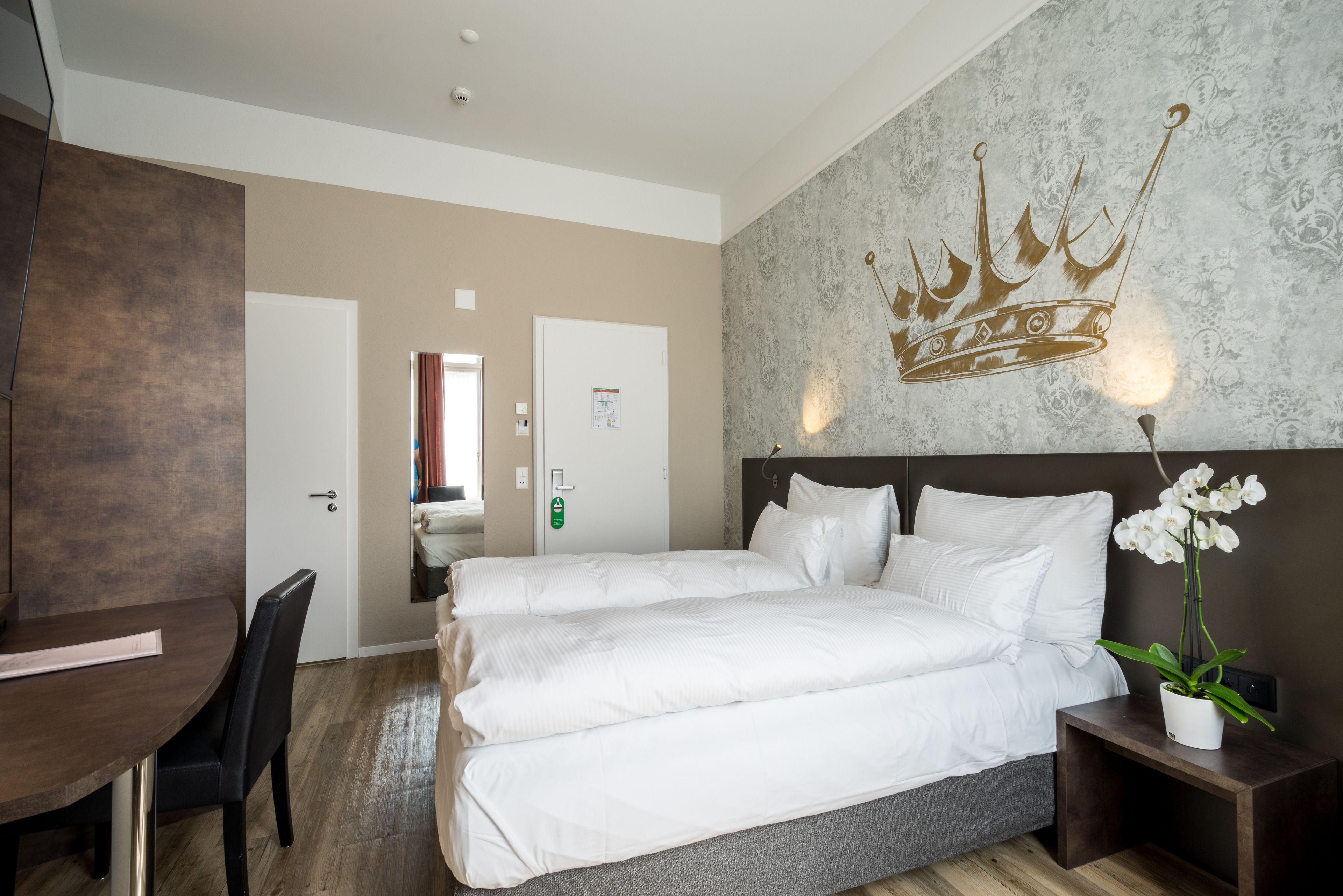 Ein Hotelzimmer mit einem großen Bett, einem Schreibtisch und einem Sessel. An der Wand ist ein Wandgemälde mit einem Kronenmotiv und auf dem Nachttisch steht ein Blumentopf.