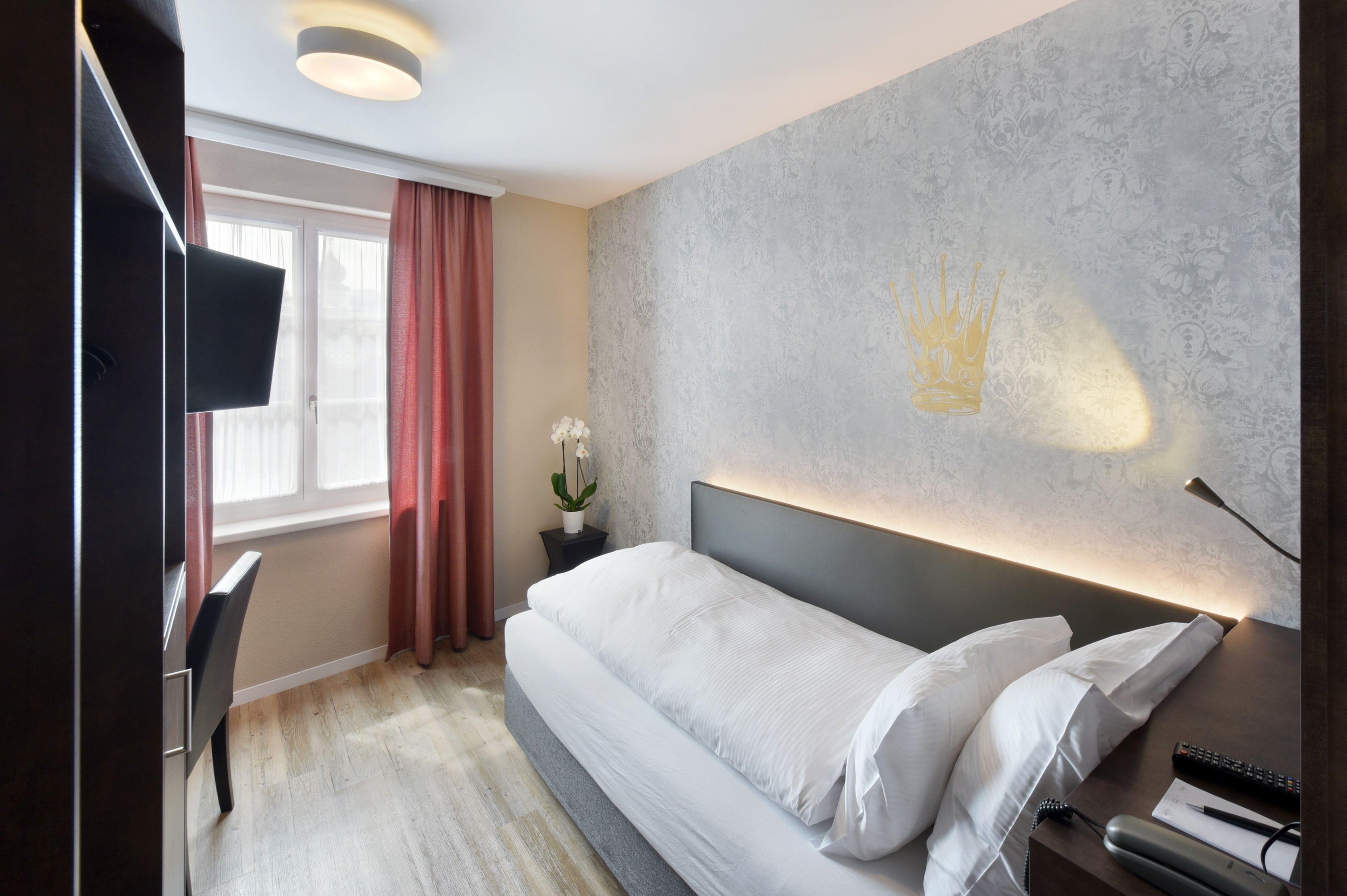 Une chambre d’hôtel comprenant un lit, un bureau, une télévision, une table de chevet et un mur orné d’un motif en forme de couronne.