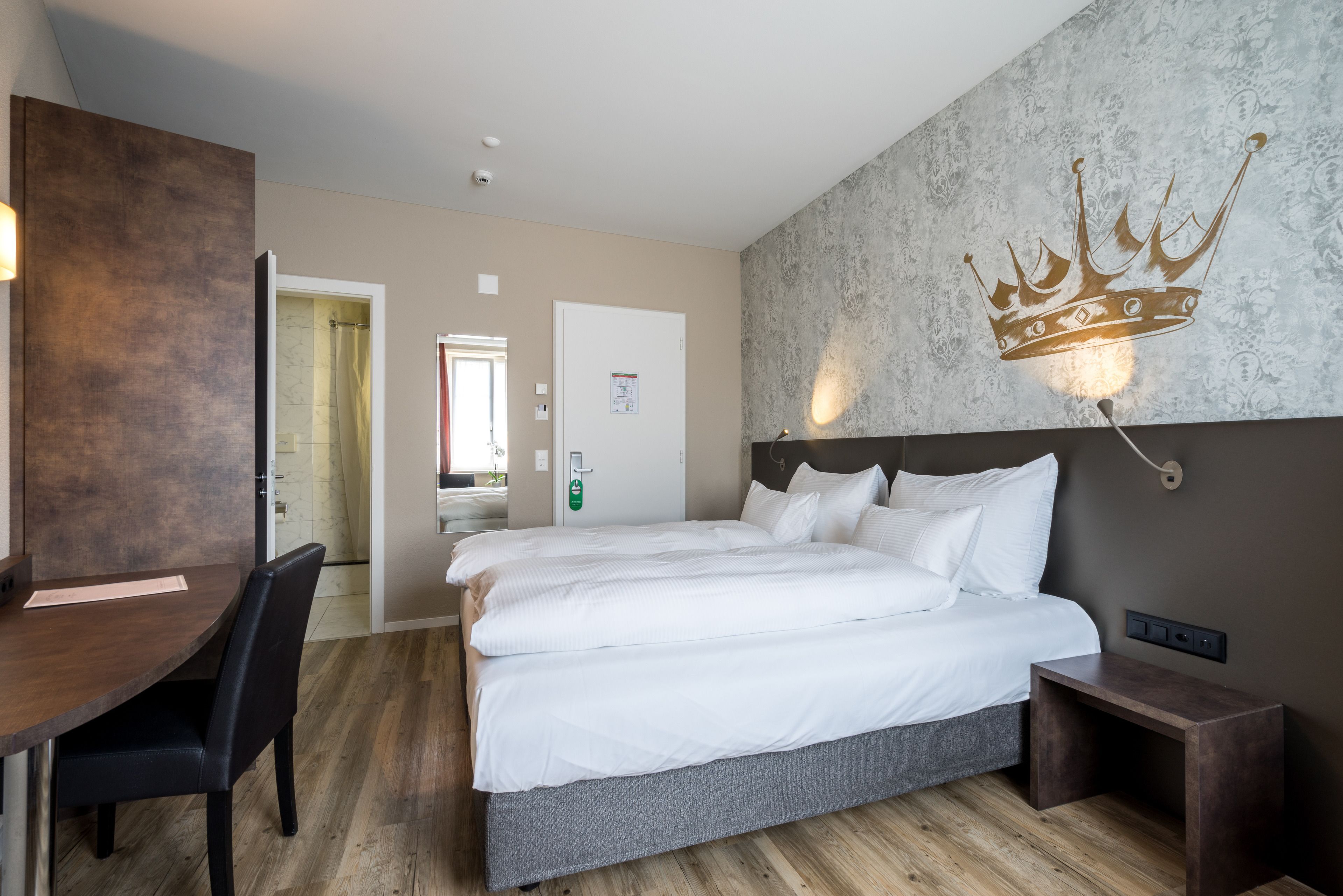 A hotel room with a large bed, a desk, and a chair. There is a crown-shaped light fixture on the wall.