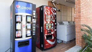 Dos máquinas expendedoras de bebidas, una de Pepsi y otra de Coca-Cola, junto a un muro de ladrillo y una zona de cocina.
