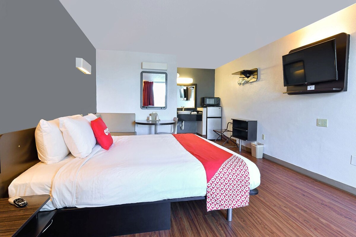 Habitación de hotel con cama, escritorio, silla, televisor, espejo y una kitchenette pequeña.