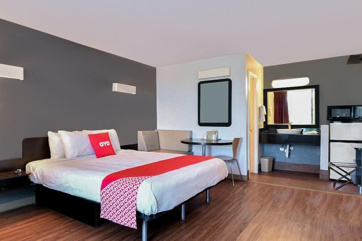 Habitación de hotel con cama, escritorio, silla, espejo, televisor y lavamanos.