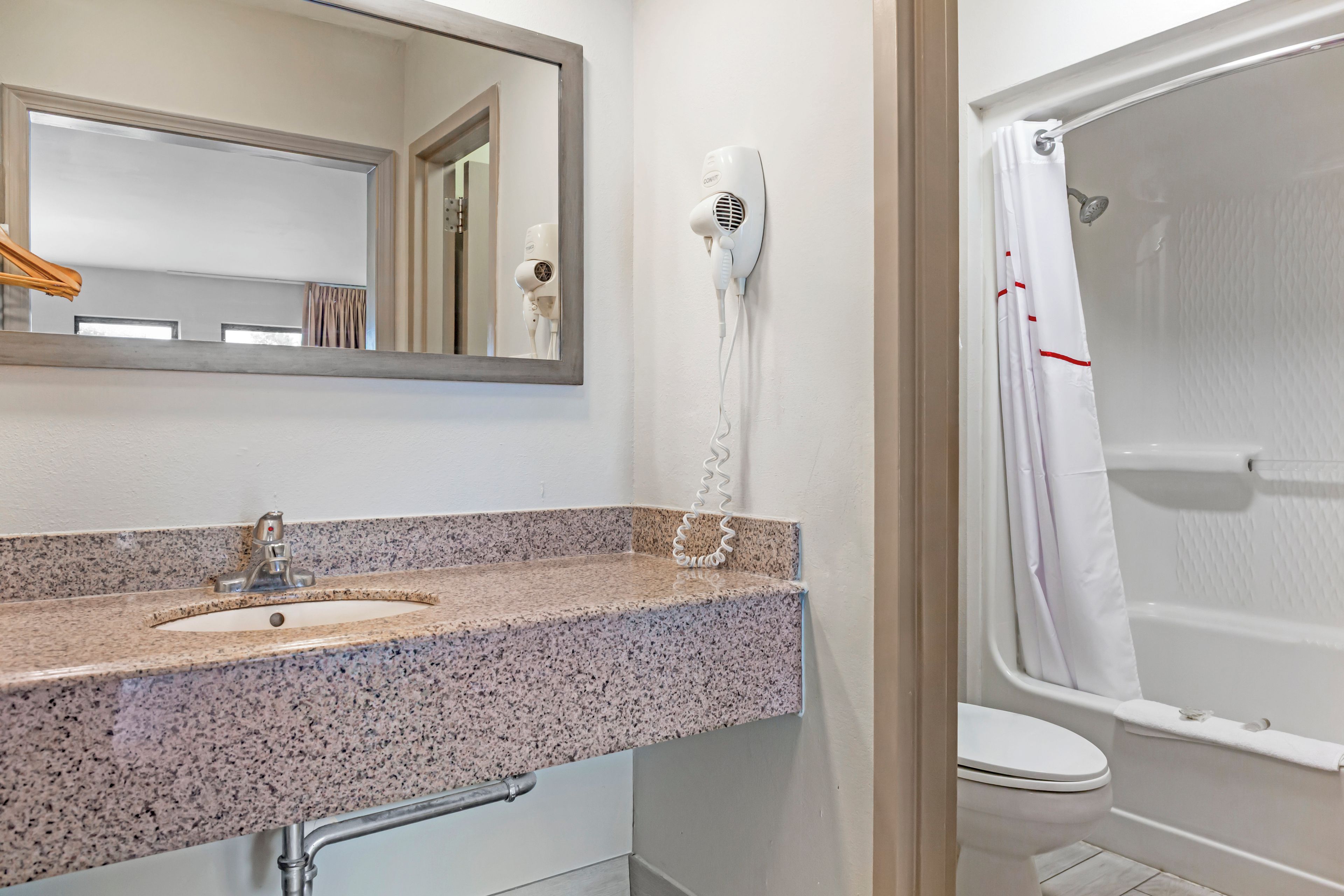 A bathroom with a granite countertop sink, a mirror, a hairdryer, and a shower with a white curtain.