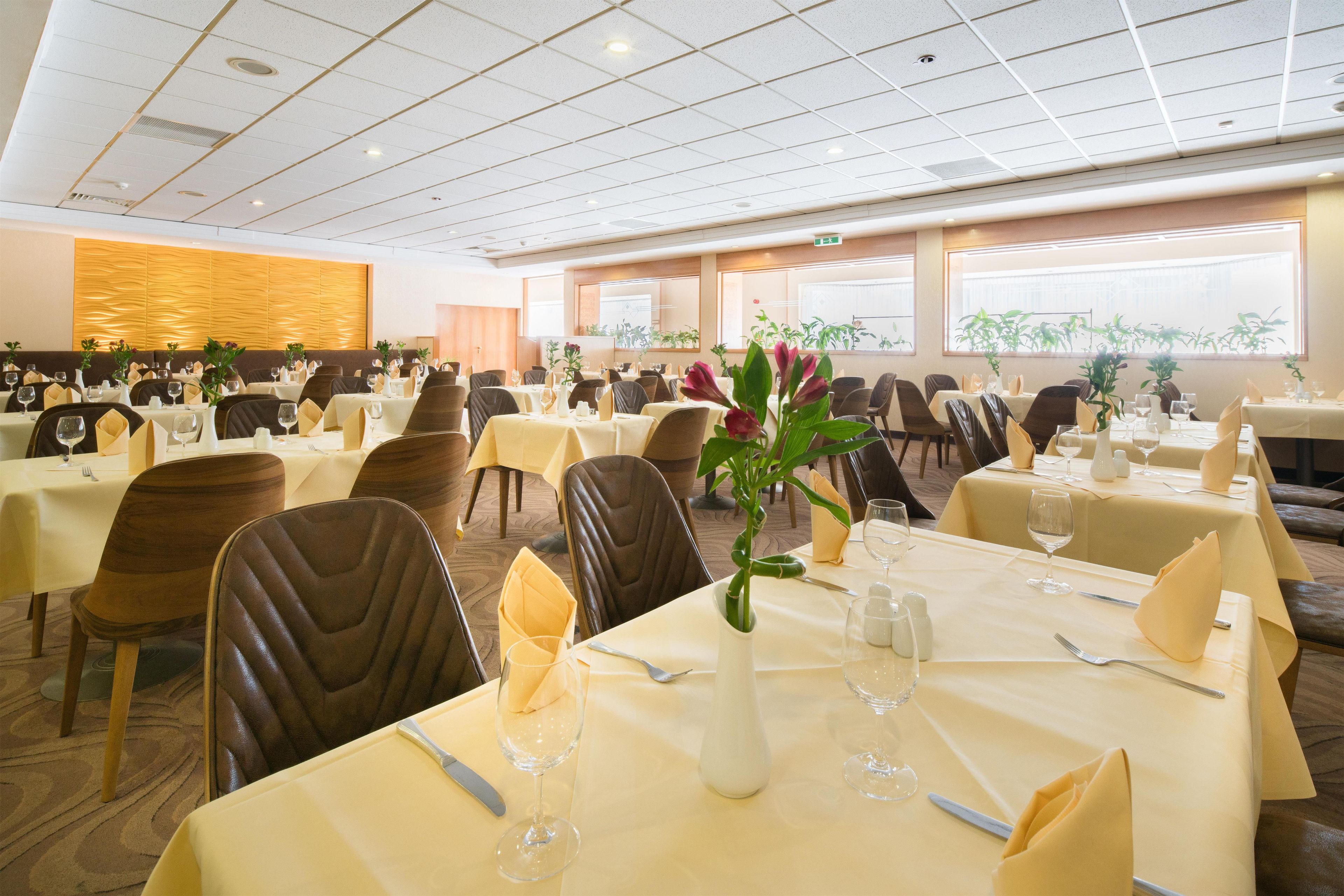 A spacious dining area with multiple tables set for guests, featuring a modern ceiling design and large windows allowing natural light.