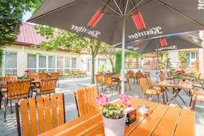 An outdoor seating area with wooden tables and chairs, covered by large umbrellas with text. - Best Western Hotel Jena (Jena)