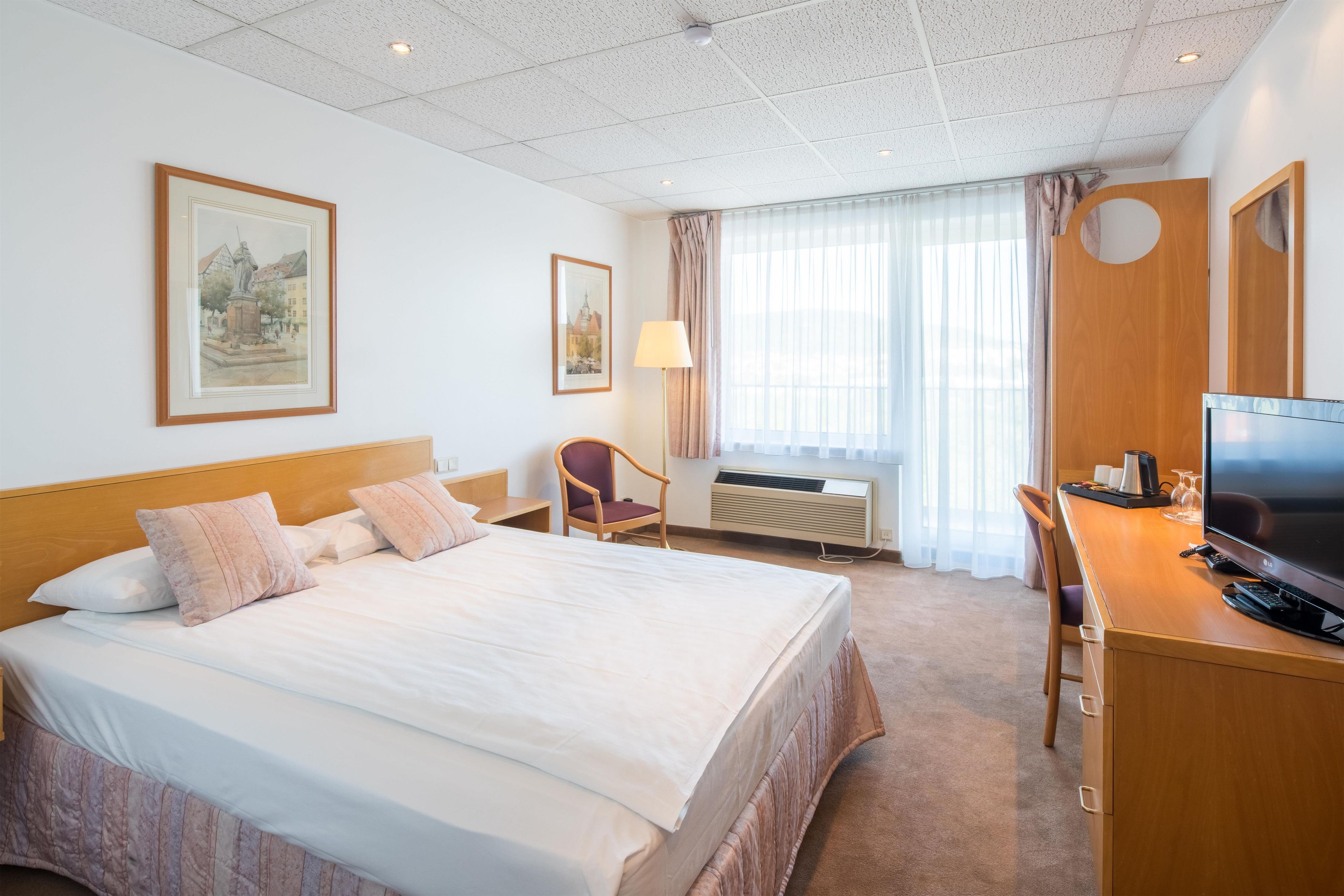 A hotel room with a large bed, a desk with a TV, a chair, and two framed pictures on the wall.
