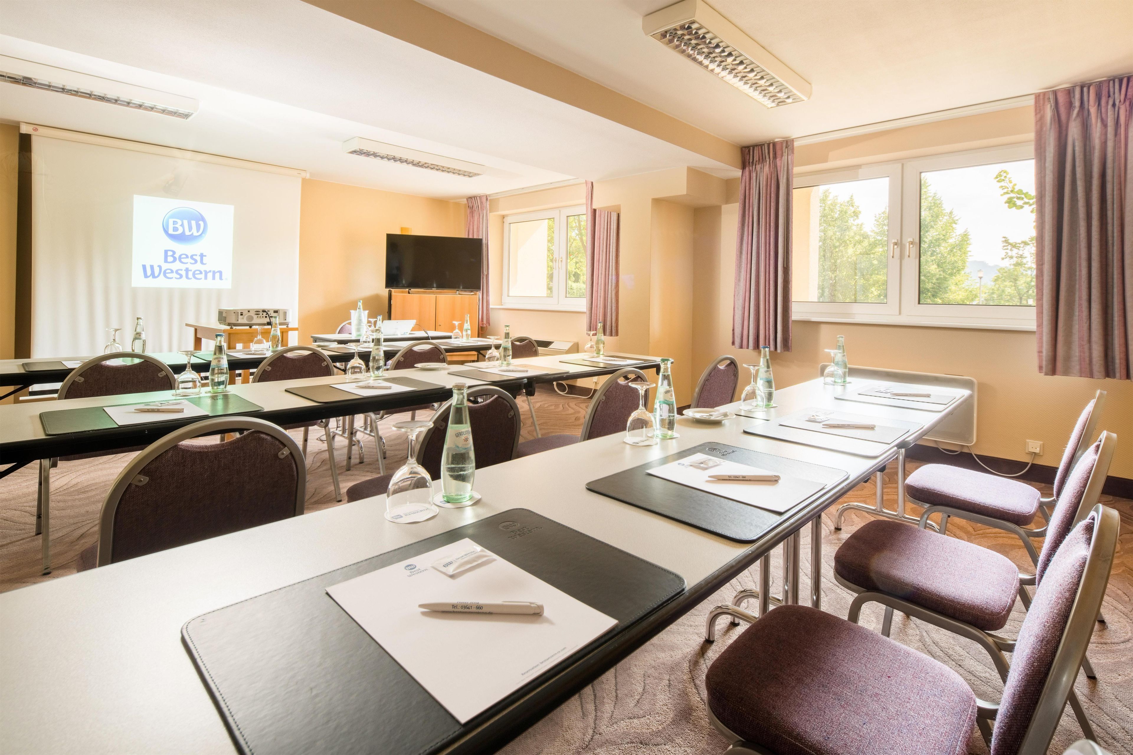 A conference room with long tables, chairs, a projector screen, and a television.